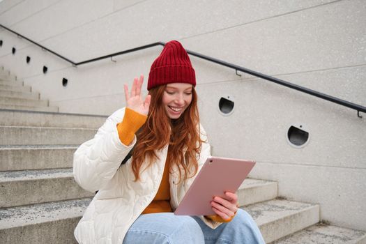 Joyful redhead teen girl, student with digital tablet, says hello, waves hand at gadget camera, connects to video chat, talks to friend in application.