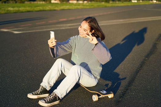 Happy asian girl sits on skateboard, takes selfie with longboard, makes cute faces, sunny day outdoors.
