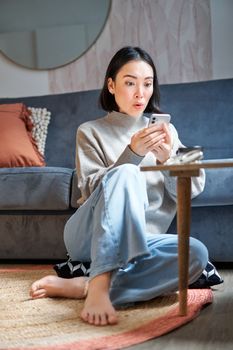 Vertical shot of young asian girl looks surprised at her mobile phone, reads amazing news, notification on smartphone app, sits at home on floor.
