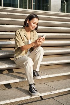 Beautiful asian girl sitting on stairs outside building, using mobile phone, looking at smartphone app and smiling.