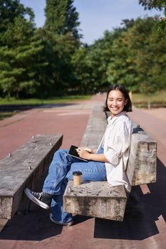 Portrait of young artist, asian girl drawing in digital tablet with graphic pen, sitting outdoors in park, getting inspiration.