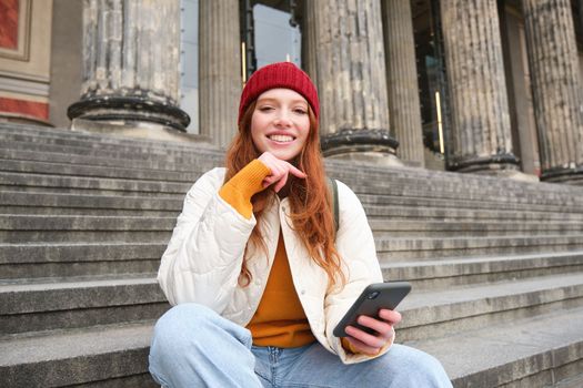 Portrait of young urban girl in red hat, sits on stairs near museum, holds mobile phone, connects to public wifi and surfs net, uses smartphone apps.