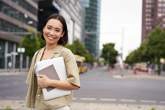 Portrait of young asian woman, looking happy and confident, going to work or university, city skyscrappers behind her, holding laptop and notebook.