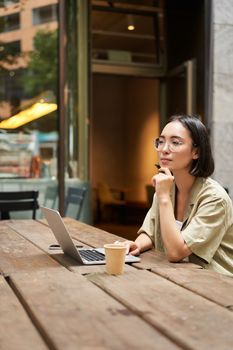 Working woman sitting with laptop in cafe and thinking. Asian girl in glasses works remotely, drinks coffee and looks thoughtful.
