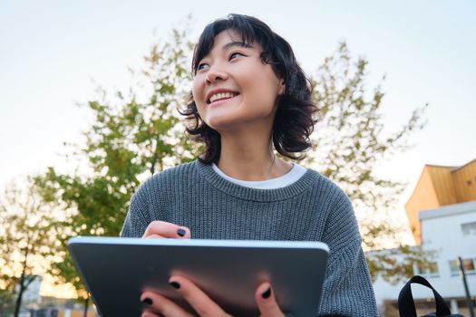 Close up portrait of young korean girl sits outdoors in park, holds her digital tablet and graphic pen, draws scatches, gets inspiration from nature for art, smiles happily.