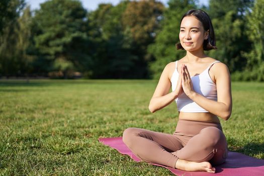 Woman meditating on lawn in park, sitting on sports mat, relaxing, breathing fresh air.