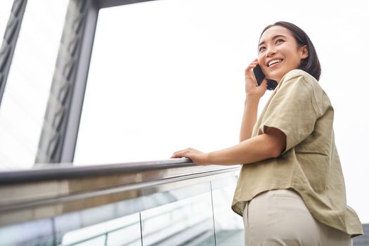 Happy asian woman on escalator, talking on mobile phone, meeting someone in city. Copy space