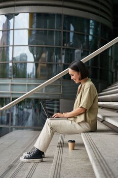 Vertical shot of smiling asian girl with coffee, uses laptop outdoors. Young woman with computer outside, sits on stairs.