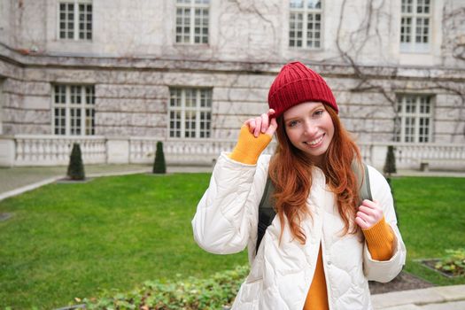 Beautiful redhead woman with backpack, tourist walking around city park and looks around, wears red hat and warm coat.