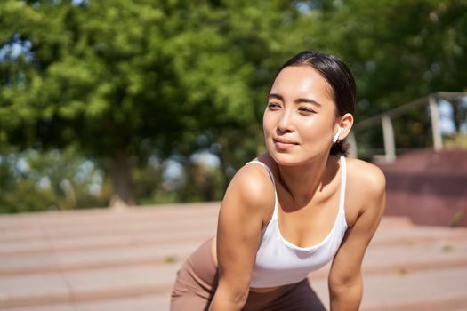 Portrait of asian woman taking break, breathing heavily and panting after running, jogger standing and wiping sweat off forehead, smiling pleased.