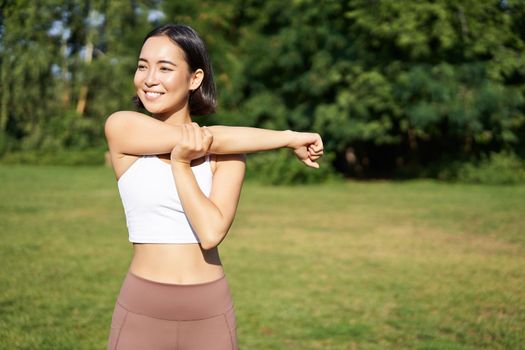 Asian woman stretching her arms, doing fitness workout in park, smiling pleased, warming up before jogging on fresh air in daytime.