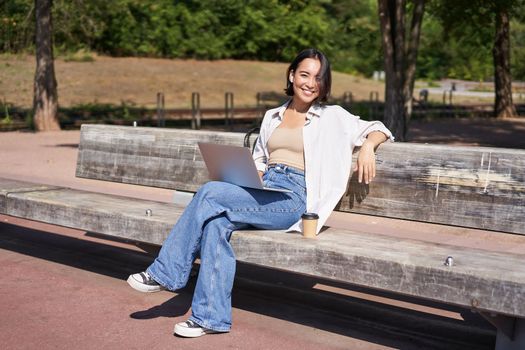 Portrait of beautiful young woman sitting on bench in park, using laptop and drinking coffee, relaxing outdoors on summer day.