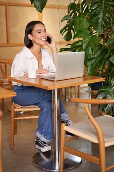 Young female cafe manager, owner sitting with laptop, answering phone calls and smiling friendly.