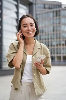 Young smiling asian woman in wireless earphones, walks on street with smartphone and headphones.