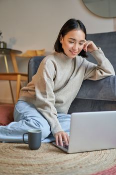 Vertical shot of young woman in cozy home working on laptop, using smartphone and drinking coffee, sitting on floor near sofa.