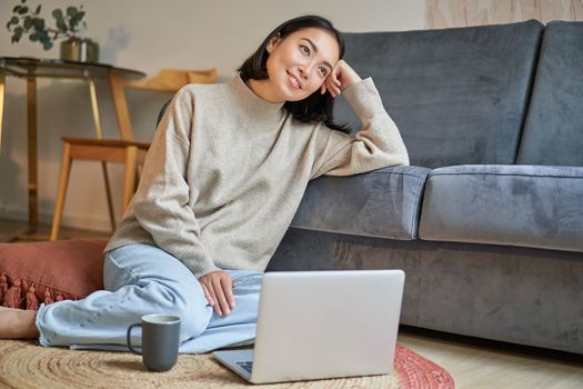Portrait of dreamy woman sitting with laptop on floor, watching on computer and drinking coffee, enjoying cozy days at home.