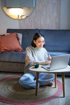 Vertical shot of asian girl sits on floor at home, working on laptop, studying at cozy place, using computer to freelance.