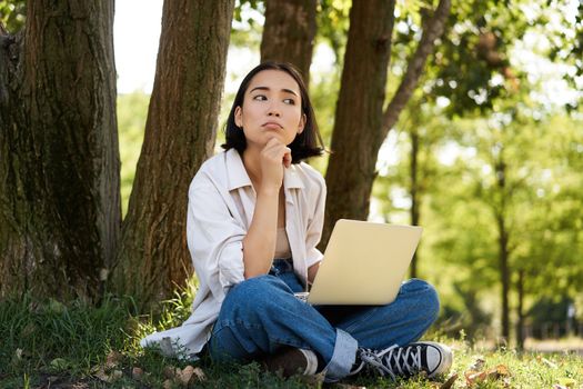 Portrait of young asian woman sitting in park near tree, working on laptop, using computer outdoors.