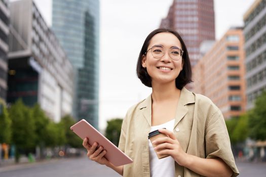 Portrait of happy young woman in glasses, standing on street with cup of coffee and tablet.