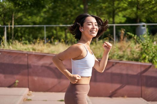 Wellbeing. Smiling asian fitness girl, runner in park, smiling and running, doing jogging workout outdoors on streets.