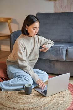 Vertical shot of young woman in cozy home working on laptop, using smartphone and drinking coffee, sitting on floor near sofa.