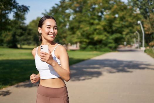 Happy smiling asian woman jogging in park. Healthy young female runner doing workout outdoors, running on streets.