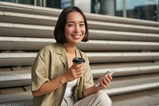 Portrait of asian woman with smartphone, drinks coffee and watches videos on mobile phone. Girl with telephone sits on stairs outdoors.