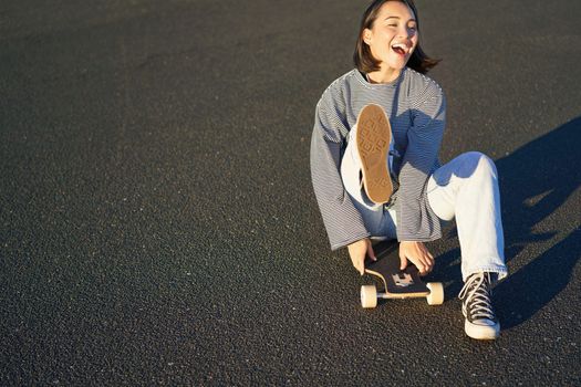 Portrait of beautiful asian girl skating, sitting on her skateboard and smiling. Cute teenager with longboard, skating on road.