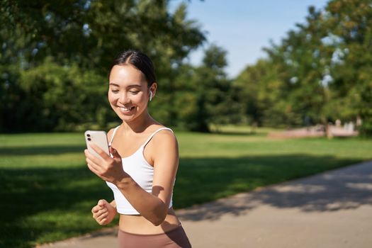 Happy asian fitness girl running and checking her stats, daily goals on smartphone app. Young woman jogging and looking at mobile screen.