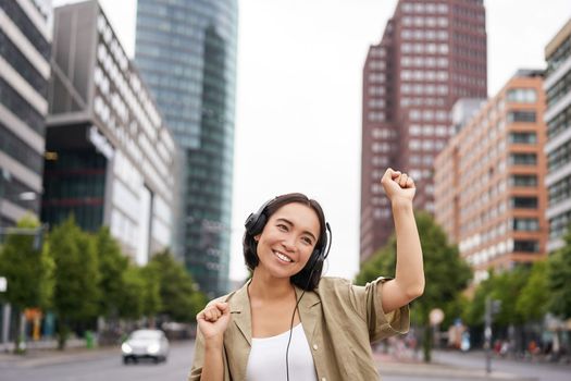 Portrait of smiling asian woman dancing, triumphing, feeling happy while listening music in city, posing on street near skyscrappers.