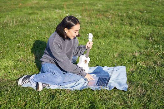 Happy smiling asian girl enjoying spring time, playing ukulele in park, looking for chords online on laptop, learning new song.