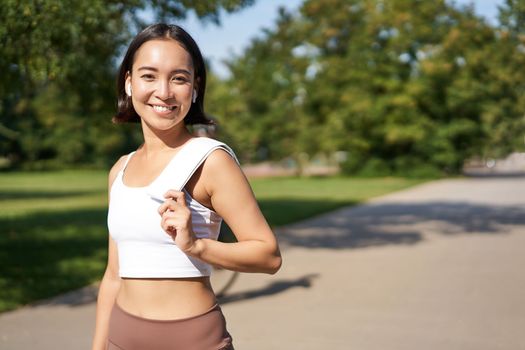 Smiling asian fitness girl holding towel on shoulder, workout in park, sweating after training exercises outdoors.
