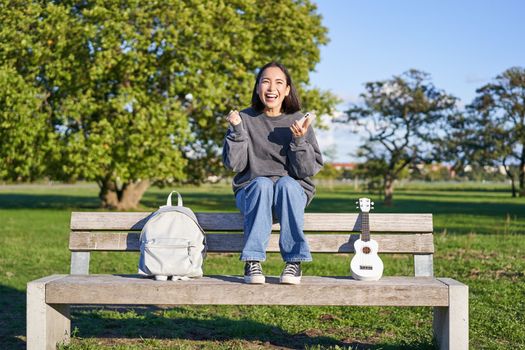 Excited girl looks at smartphone and celebrates, wins on mobile phone, sits with ukulele and backpack in park on bench on sunny day.