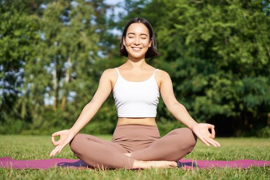 Sporty woman meditating on fresh air, sitting on fitness mat and practice yoga, smiling pleased. Sport and people concept.