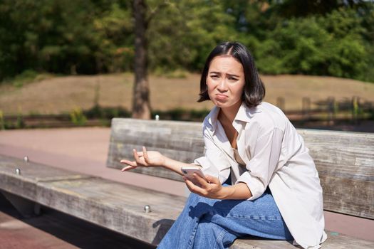Depressed asian girl sits on bench in park with smartphone, feeling uneasy and stressed, frowning and sighing.