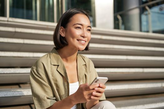 People and technology. Smiling beautiful asian woman sitting on stairs in city, holding mobile phone. Girl with smartphone rests outside.