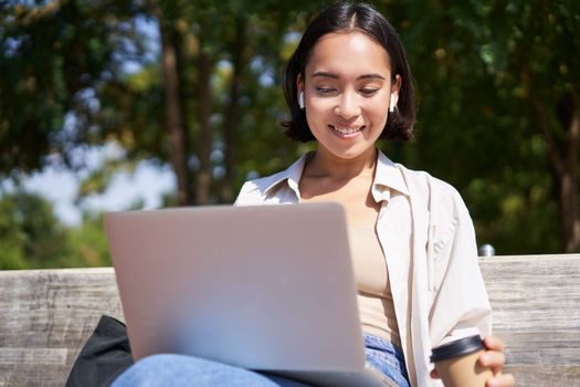 Asian girl sitting with laptop and wireless earphones, drinking coffee, looking at screen, doing homework, working on remote.