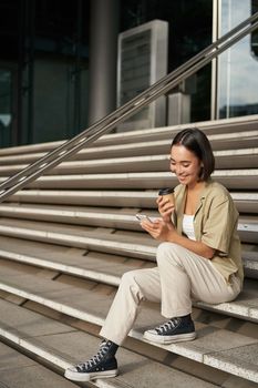 Vertical shot of young smiling woman sits on stairs in city centre wth mobile phone. Girl with telephone, browsing on smartphone, texting message.