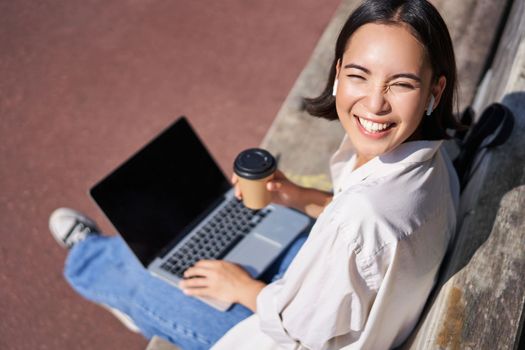 Beautiful young asian female student, sitting with laptop on bench in park, drinking takeaway coffee and listening music, working remotely, studying outdoors.