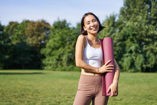 Portrait of asian girl smiles and laughs, stands with sports equipment, rubber mat, wears uniform for workout and exercises in park.