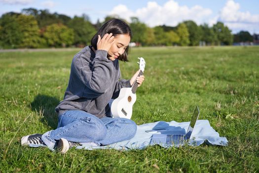 Smiling asian girl learns how to play ukulele via laptop, online video tutorials, sitting on grass in park with musical instrument.