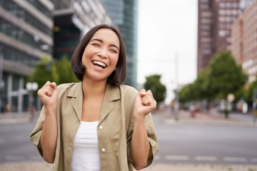 Happy asian girl triumphing on streets of city, dancing from happiness, celebrating victory, posing outdoors.