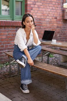 Vertical shot of trendy asian girl sitting in cafe alone with laptop, working or studying, browsing on computer. People and lifestyle concept