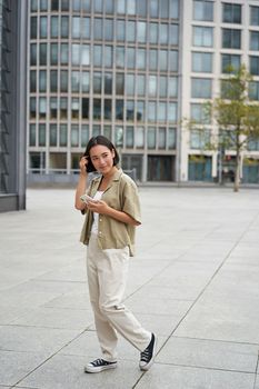 Portrait of asian woman walking in city. Street style shot of girl with smartphone, posing outdoors on street.