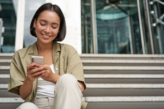 Smiling asian girl sits on stairs near building entrance, using mobile phone app. Happy young woman rests with smartphone in her hands outdoors.