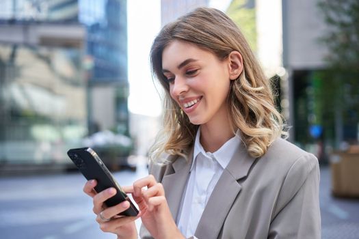 Smiling young businesswoman using mobile phone while standing outdoors. Corporate people concept.