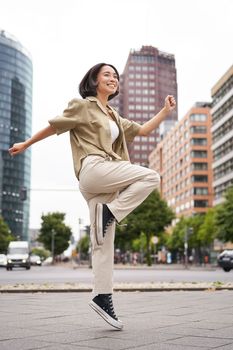 Vertical shot of young asian woman posing happy, raising hands up and dancing, triumphing, celebrating victory, enjoying day out in city.