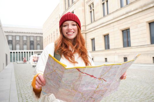 Beautiful redhead woman, tourist with city map, explores sightseeing historical landmark, walking around old town, smiling happily.