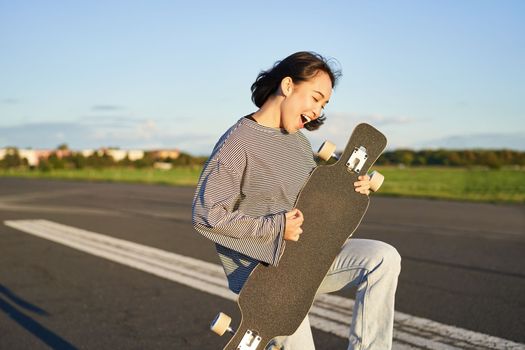 Beautiful asian teen girl playing with her longboard, holding skateboard as if playing guitar, standing on road on sunny day.