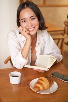 Vertical shot of happy young asian woman enjoys reading, sitting with book in cafe, drinking coffee and eating croissant.